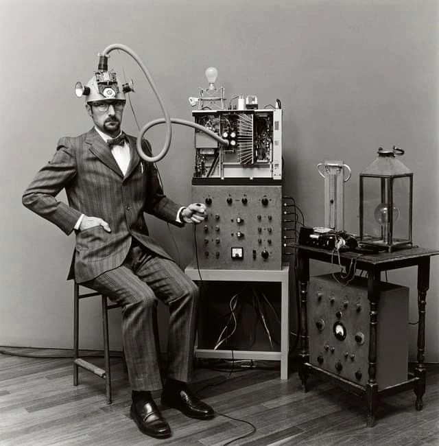 A seated inventor beside an experimental apparatus in a historical visual reference.