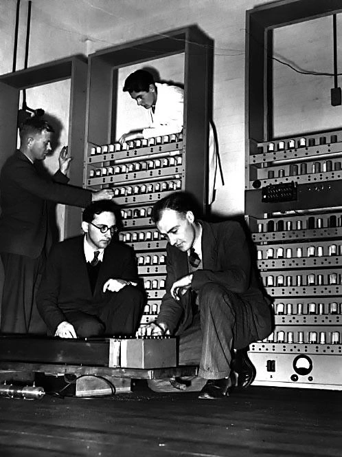 Researchers working together among tall racks of early computing equipment.