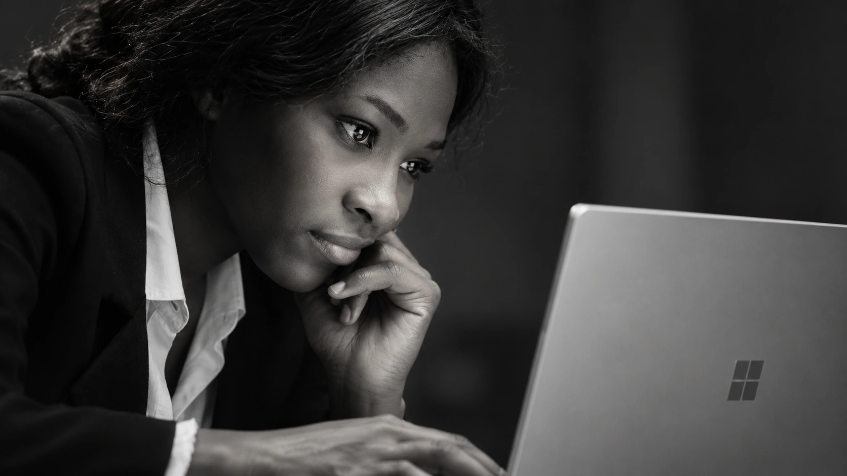 A cinematic black and white close shot of a woman studying her Windows laptop, her face illuminated by its screen.