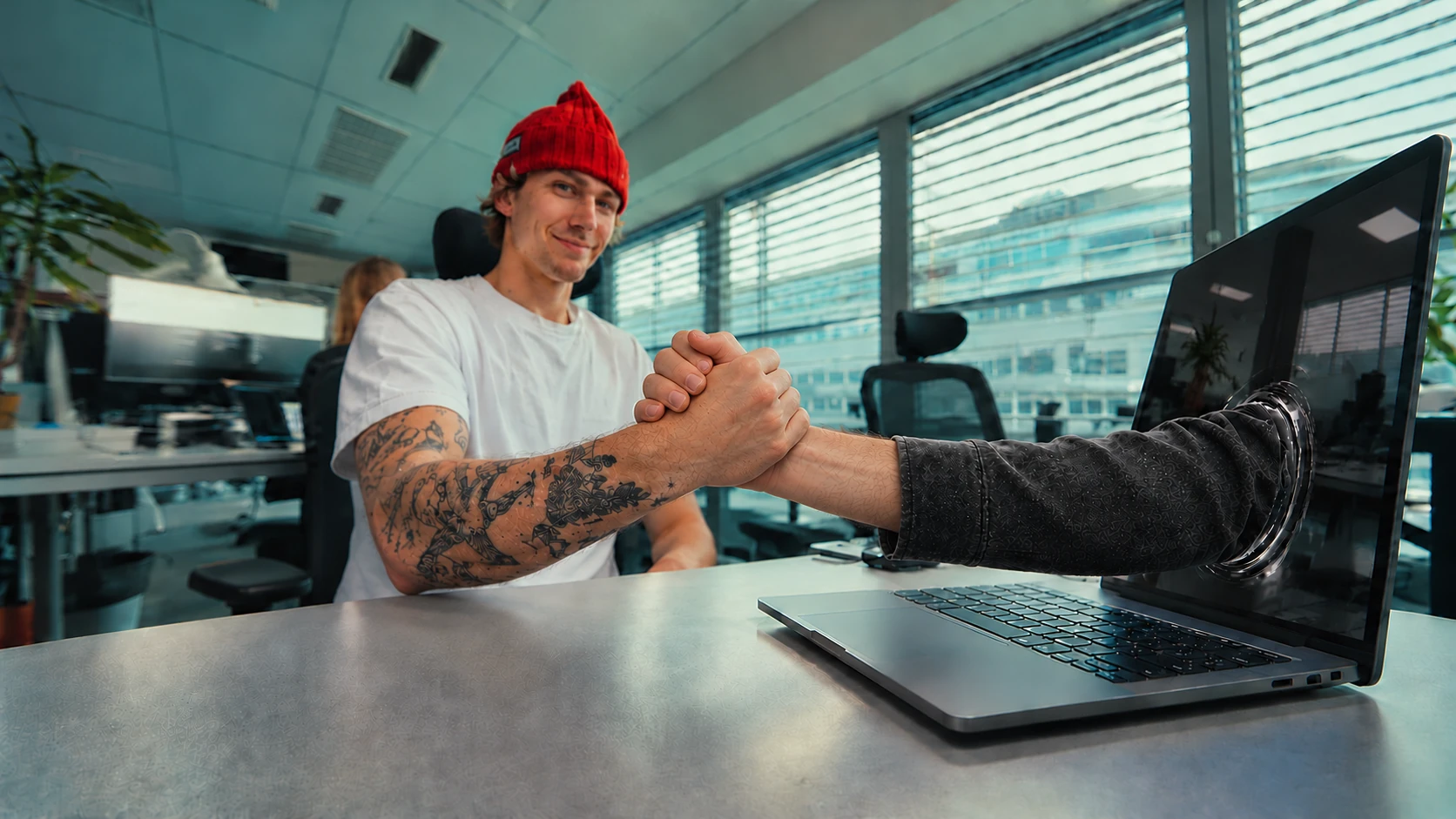 A designer in a red beanie greets the hand reaching through his laptop screen.